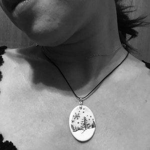 May include: Oval pendant necklace with a winter scene design. The pendant features a black and white illustration of a snowy landscape with trees and snowflakes. The pendant hangs from a black cord necklace.