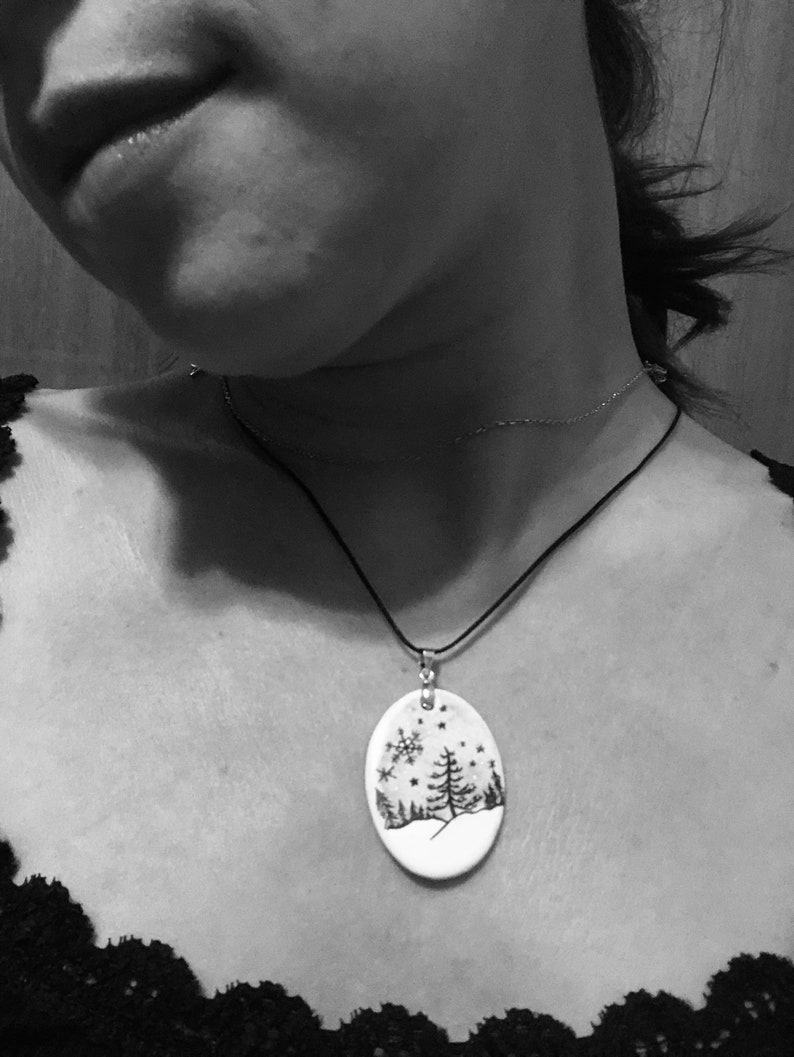 May include: Oval pendant necklace with a winter scene design. The pendant features a black and white illustration of a snowy landscape with trees and snowflakes. The pendant hangs from a black cord necklace.