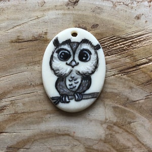May include: Oval pendant with a hand-painted owl design. The owl is in black and white, perched on a branch. The pendant has a small hole at the top for stringing. The background is a wooden surface.