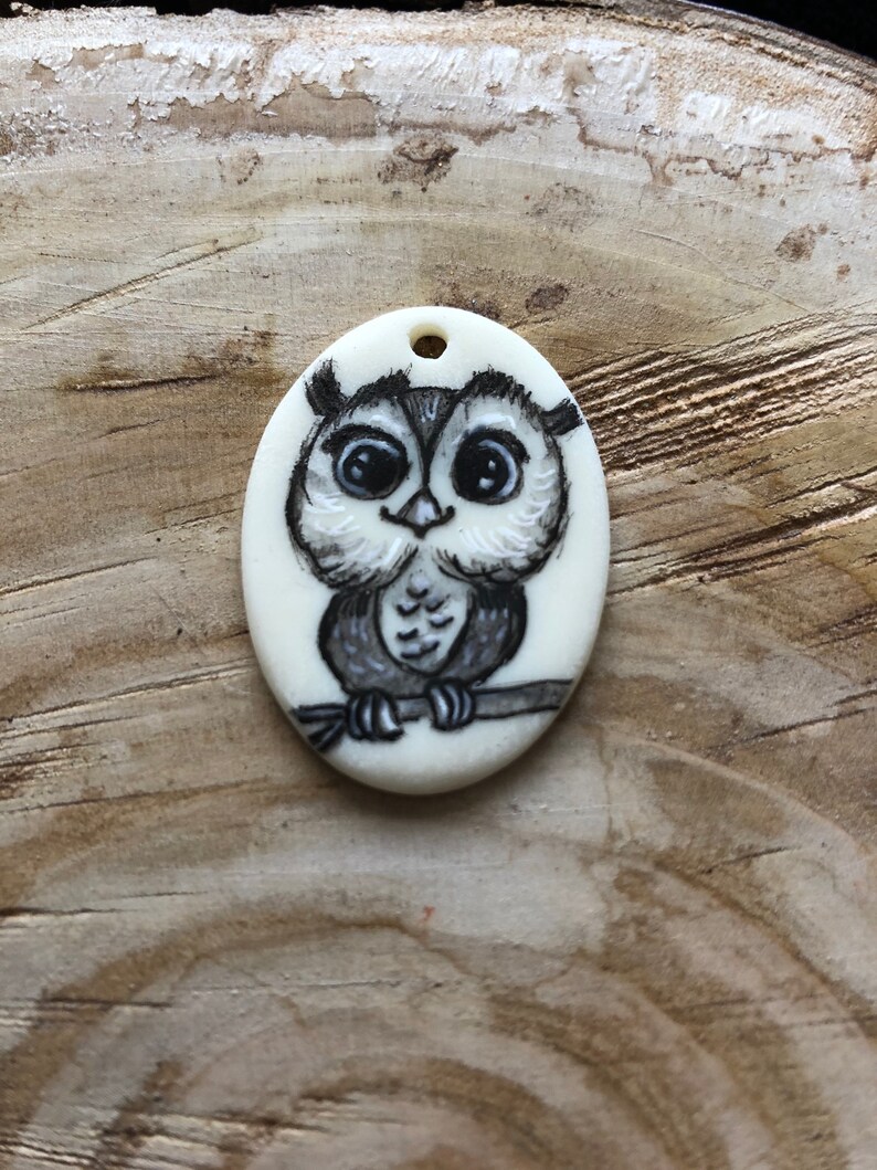 May include: Oval pendant with a hand-painted owl design. The owl is in black and white, perched on a branch. The pendant has a small hole at the top for stringing. The background is a wooden surface.