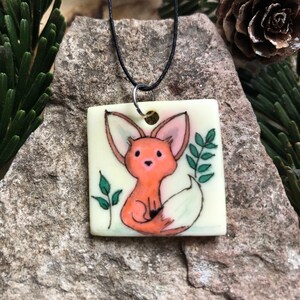 May include: Square pendant necklace with a hand-painted fox illustration. The fox is orange with pink ears, set against a cream background. Green leaves and a black cord complete the design. The pendant measures approximately 3.8 cm square.