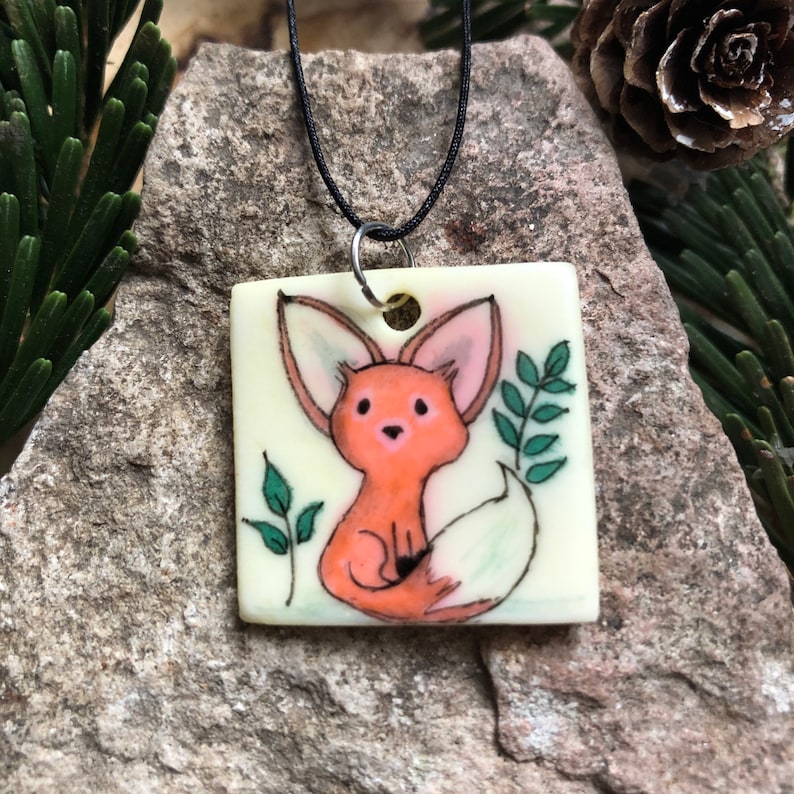 May include: Square pendant necklace with a hand-painted fox illustration. The fox is orange with pink ears, set against a cream background. Green leaves and a black cord complete the design. The pendant measures approximately 3.8 cm square.