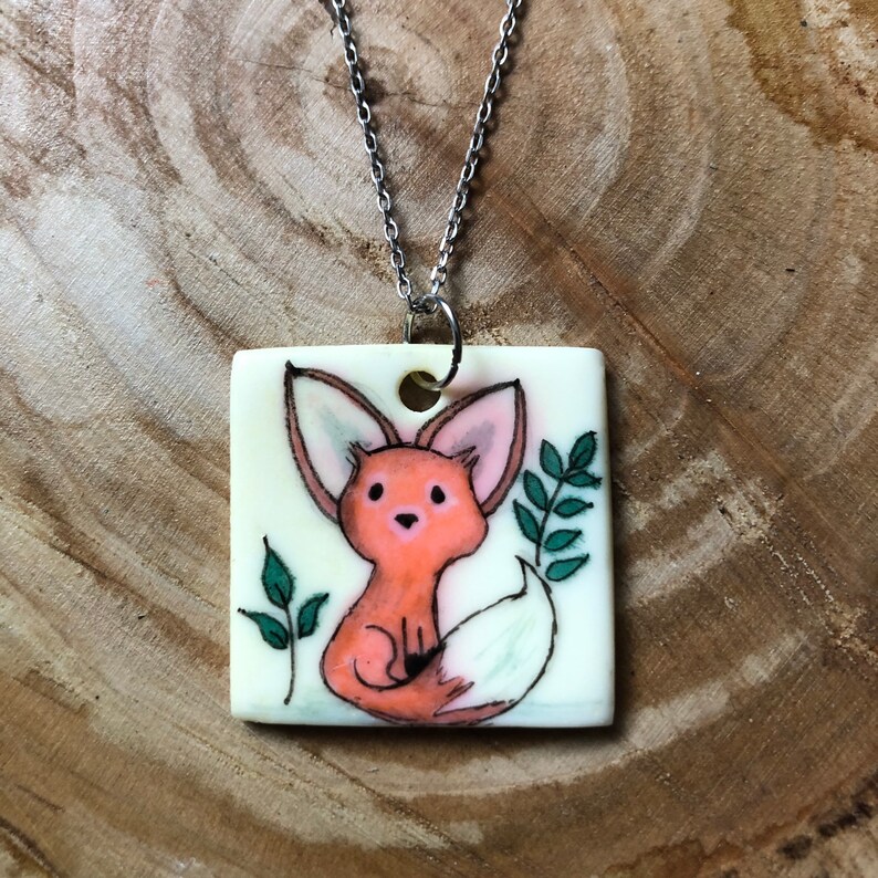May include: A square pendant necklace featuring a hand-drawn fox illustration. The fox is orange with a white tail, set against a cream background. Green leaves flank the fox. The pendant is suspended from a silver chain.