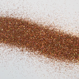 Op de afbeelding: Een strooisel van koperkleurige glitter op een witte achtergrond. De glitter is geconcentreerd in een lange, onregelmatige vorm, met individuele deeltjes die licht reflecteren. Het algehele effect is een glinsterend, getextureerd oppervlak.