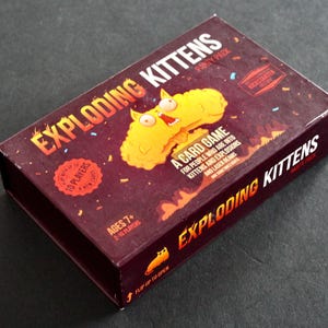 Peut inclure: Bo&icirc;te rectangulaire rouge fonc&eacute; pour le jeu de cartes "Exploding Kittens Party Pack". La bo&icirc;te pr&eacute;sente le titre du jeu en orange et blanc, et une image de chat jaune. Le texte comprend "Ages 7+" et "10 joueurs".