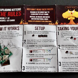 Peut inclure: Les r&egrave;gles d'Exploding Kittens, un jeu de cartes, sont affich&eacute;es sur une feuille pli&eacute;e. La feuille comprend des instructions sur la fa&ccedil;on de jouer, la configuration et les tours. Un petit appareil &eacute;lectronique est visible dans le coin.
