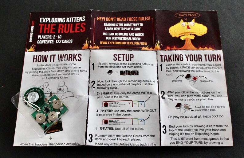 Peut inclure: Les r&egrave;gles d'Exploding Kittens, un jeu de cartes, sont affich&eacute;es sur une feuille pli&eacute;e. La feuille comprend des instructions sur la fa&ccedil;on de jouer, la configuration et les tours. Un petit appareil &eacute;lectronique est visible dans le coin.