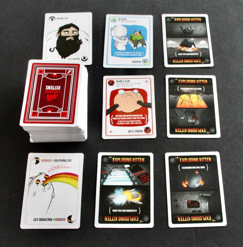 Peut inclure: Jeu de cartes "Exploding Kittens" avec des illustrations humoristiques. On distingue des cartes avec des dessins de personnages et des phrases. Une pile de cartes avec un motif rouge et blanc est &eacute;galement pr&eacute;sente.