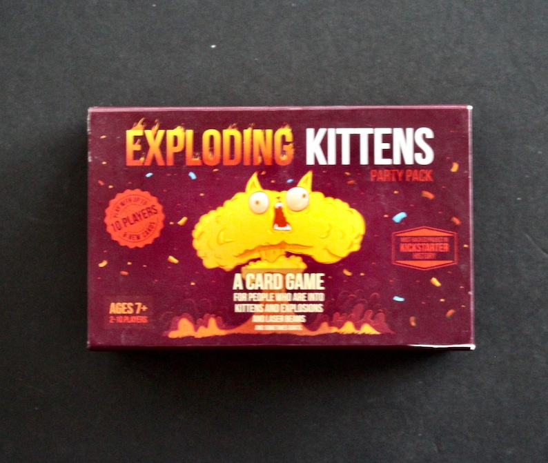 Peut inclure: Une bo&icirc;te rectangulaire pour le jeu de cartes "Exploding Kittens". La bo&icirc;te est bordeaux avec le titre du jeu en orange et une explosion de dessin anim&eacute; avec un visage de chat. Le texte sur la bo&icirc;te comprend "Party Pack", "10 joueurs" et "&Acirc;ges 7+".