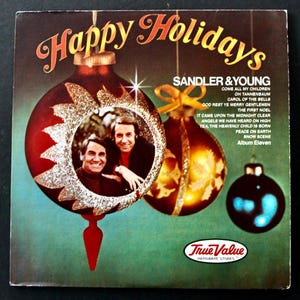 May include: Vintage vinyl record album cover featuring the text "Happy Holidays" in gold script. The cover art includes images of Christmas ornaments and a photo of Sandler & Young. The album title is "Album Eleven".