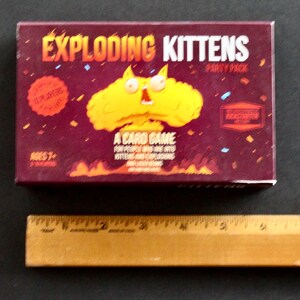 Peut inclure: Bo&icirc;te rectangulaire du jeu de cartes "Exploding Kittens Party Pack". La bo&icirc;te bordeaux pr&eacute;sente du texte et des graphiques orange et jaune. Le jeu est pour 2 &agrave; 10 joueurs, &agrave; partir de 7 ans. La bo&icirc;te pr&eacute;sente une explosion de dessin anim&eacute; avec un visage de chat.