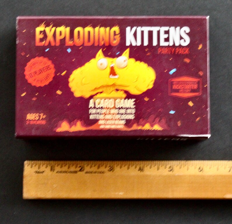 Peut inclure: Bo&icirc;te rectangulaire du jeu de cartes "Exploding Kittens Party Pack". La bo&icirc;te bordeaux pr&eacute;sente du texte et des graphiques orange et jaune. Le jeu est pour 2 &agrave; 10 joueurs, &agrave; partir de 7 ans. La bo&icirc;te pr&eacute;sente une explosion de dessin anim&eacute; avec un visage de chat.