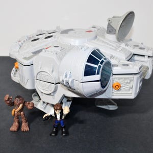 May include: A light gray plastic spaceship toy with a circular radar dish, a cockpit with dark blue windows, and intricate molded surface details. Two small plastic figures, one brown and furry and one human-like, stand in front of the ship.