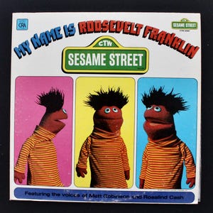 May include: Vintage Sesame Street record album cover titled "My Name is Roosevelt Franklin." The cover features three images of the character Roosevelt Franklin in a striped shirt and a dark afro, set against pink, yellow, and blue backgrounds.