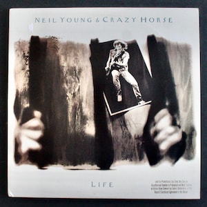 May include: Album cover for "Life" by Neil Young & Crazy Horse. The cover features a black and white image of a man playing a guitar, set against an abstract background. The band's name is printed at the top, and the album title is at the bottom.