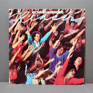 May include: Album cover for Richard Simmons' "Reach" featuring a crowd with arms raised. The title is in a light blue script. The crowd is diverse, with people wearing various colors. The album cover has a vintage aesthetic.