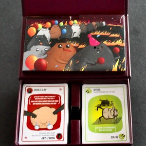 Peut inclure: Un coffret bordeaux contenant un jeu de cartes. Le couvercle de la bo&icirc;te pr&eacute;sente une illustration color&eacute;e de personnages de dessins anim&eacute;s lors d'une f&ecirc;te avec des ballons. &Agrave; l'int&eacute;rieur, deux jeux de cartes sont visibles, l'un intitul&eacute; "DOUBLE SLAP" et l'autre "DEFUSE".