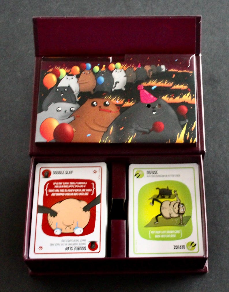 Peut inclure: Un coffret bordeaux contenant un jeu de cartes. Le couvercle de la bo&icirc;te pr&eacute;sente une illustration color&eacute;e de personnages de dessins anim&eacute;s lors d'une f&ecirc;te avec des ballons. &Agrave; l'int&eacute;rieur, deux jeux de cartes sont visibles, l'un intitul&eacute; "DOUBLE SLAP" et l'autre "DEFUSE".