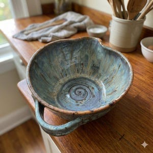 May include: A handmade ceramic bowl featuring a mottled blue and grey glaze with a spiral pattern at the base. The bowl has an irregular, organic rim and a single sturdy handle extending from one side, showcasing a rustic, textured finish throughout.