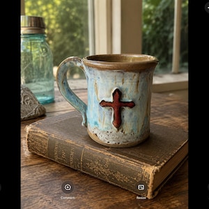 May include: A ceramic mug with a light blue and cream glaze featuring a raised, dark red cross emblem on the front. The mug has a sturdy handle and a wide rim, with visible brown clay tones at the base and top edge, showcasing a handcrafted texture.