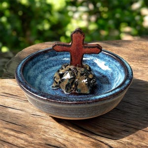 May include: A handmade ceramic bowl with a dark blue glaze and a raised, textured base. A reddish-brown cross is mounted on a dark, textured ceramic base in the center of the bowl. The bowl is likely used for religious purposes.