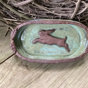 May include: A green ceramic oval dish with a brown rabbit design. The dish has a brown rim with a braided design.