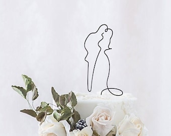 May include: A white tiered wedding cake adorned with cascading ivory roses, hydrangeas, and greenery. A delicate black wire sculpture of a couple kissing tops the cake. The cake is a centerpiece for a wedding celebration.
