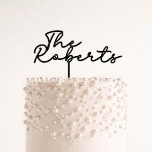 May include: A black cake topper reading "The Roberts" in a cursive script. The topper sits atop a white cake, adorned with white, pearl-like decorations, creating an elegant design.