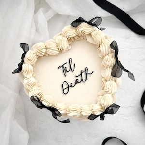 May include: A heart-shaped cake with cream-colored frosting and the words "Til Death" in black script. The cake is decorated with black ribbon bows and sits on a white surface.