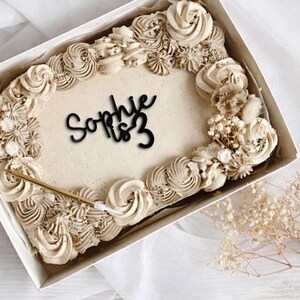 May include: A rectangular cake in a white box, decorated with beige frosting swirls and floral accents. The cake has the name "Sophie" and the number "3" in black script. A gold candle is inserted into the cake.