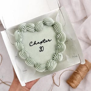 May include: A heart-shaped cake with sage green frosting and the words "Chapter 30" in black script. The cake is in a white box, with a roll of twine visible.
