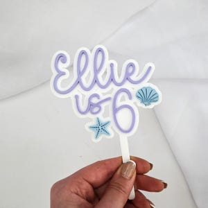 May include: A white and lavender cake topper with the text "Ellie is 6". The topper features a seashell and starfish design. The topper is held by a white stick.