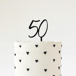 May include: A black '50' cake topper on a white cake decorated with black heart sprinkles. The cake has a textured surface and is set against a white background. Ideal for a 50th birthday celebration.