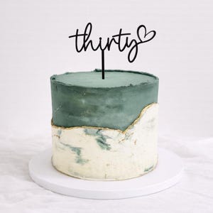 May include: A two-tiered cake with a teal-green top and a marbled white and green bottom. The cake is decorated with a gold trim and a black cake topper that reads "thirty" with a heart symbol.