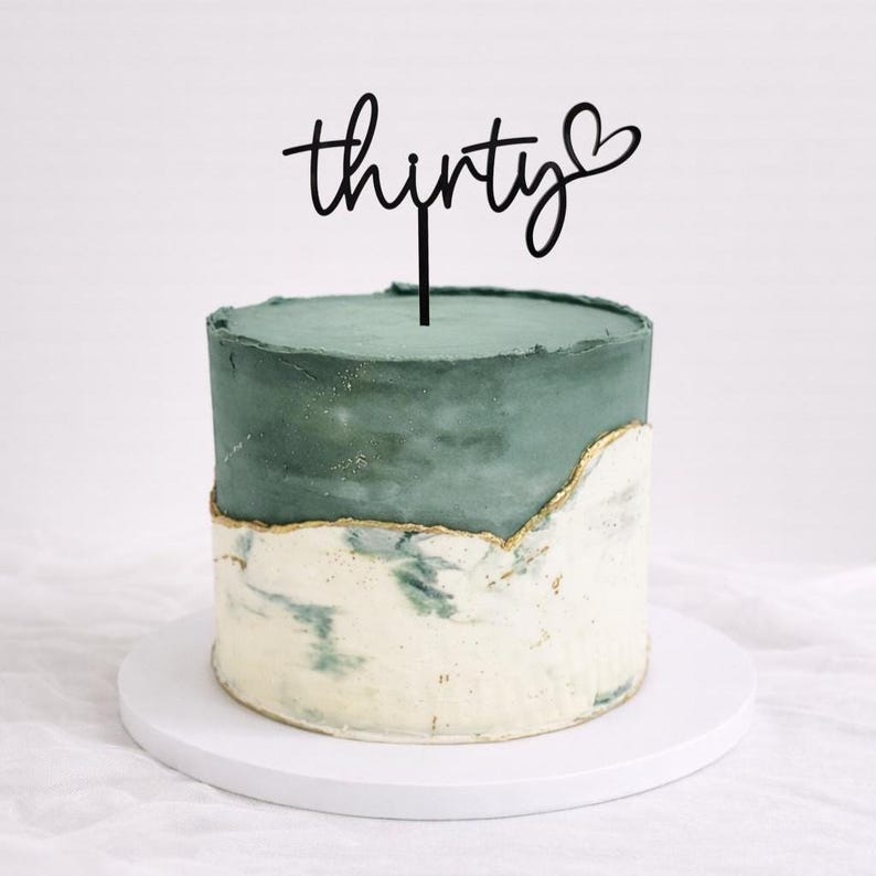 May include: A two-tiered cake with a teal-green top and a marbled white and green bottom. The cake is decorated with a gold trim and a black cake topper that reads "thirty" with a heart symbol.