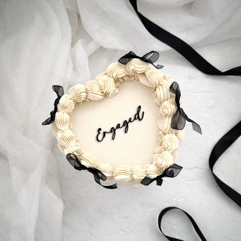 Puede incluir: Un pastel en forma de coraz&oacute;n con glaseado color crema y la palabra "Engaged" en escritura negra. El pastel est&aacute; decorado con glaseado color crema en los bordes y lazos de cinta negra. El pastel est&aacute; sobre una superficie blanca con cinta negra.