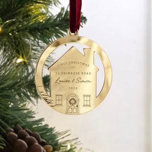 May include: A circular gold-toned metal Christmas ornament featuring a house-shaped cutout. The surface is engraved with the text "FIRST CHRISTMAS AT 55 PRIMROSE ROAD", "Louise & Simon", and "2026". It hangs from a dark red velvet ribbon.