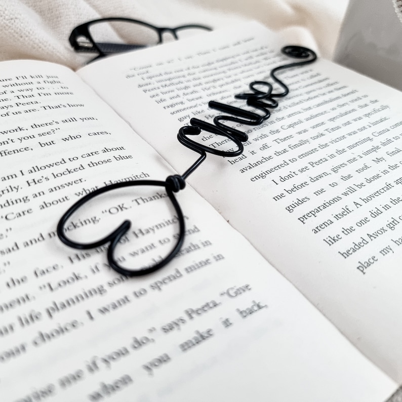 May include: A black wire bookmark in the shape of the word "Chloe" with a heart design. The bookmark rests on the open pages of a book. A pair of black glasses is in the background.
