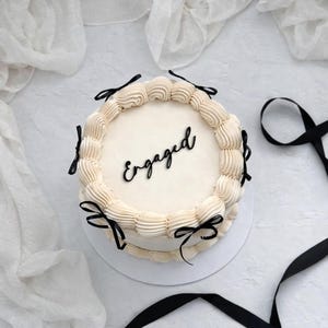 Puede incluir: Una tarta redonda con glaseado blanco y la palabra "Engaged" en escritura negra. La tarta est&aacute; decorada con remolinos de glaseado color crema y lazos de cinta negra. La tarta est&aacute; sobre una superficie blanca.