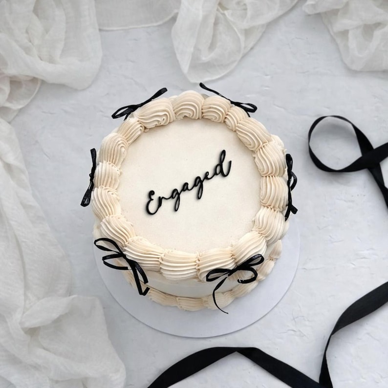 Puede incluir: Una tarta redonda con glaseado blanco y la palabra "Engaged" en escritura negra. La tarta est&aacute; decorada con remolinos de glaseado color crema y lazos de cinta negra. La tarta est&aacute; sobre una superficie blanca.