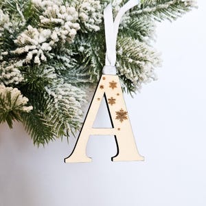 May include: A wooden letter A Christmas ornament with a white ribbon and gold snowflake accents.