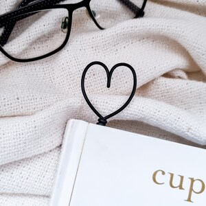 May include: A black metal heart-shaped bookmark is inserted into a white book with gold lettering. The bookmark is on a white knitted blanket. Black eyeglasses with a dotted pattern are in the background.
