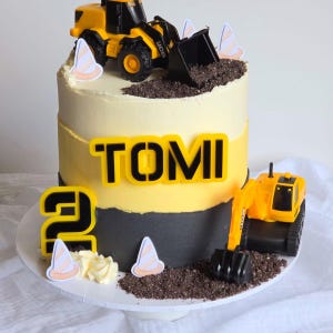 May include: A birthday cake decorated with a construction theme. The cake is yellow, black, and white, with a toy excavator on top. The name "TOMI" and the number "2" are displayed on the cake.