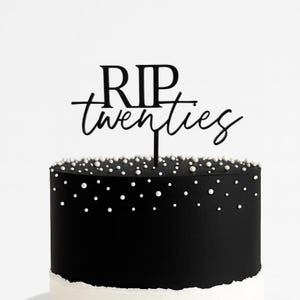 May include: A black cake with white pearl sprinkles and a white base. A black cake topper reads "RIP twenties" in a script font. The cake is set against a white background.