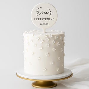 May include: A white cake decorated with small white flowers, topped with a round sign that reads "Evie's Christening" and the date 16.03.25. The cake sits on a white and gold cake stand.