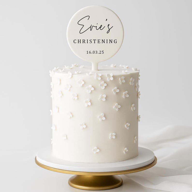 May include: A white cake decorated with small white flowers, topped with a round sign that reads "Evie's Christening" and the date 16.03.25. The cake sits on a white and gold cake stand.