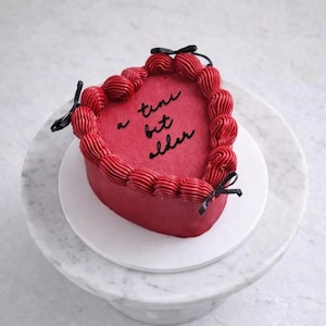 May include: A heart-shaped red cake with piped frosting around the edge and black bows. The cake has the words "a tiny bit older" written in black. The cake sits on a white plate atop a marble cake stand.
