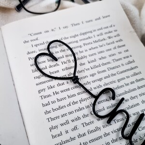 May include: A black metal heart-shaped bookmark rests on the open pages of a book. The bookmark is made of a single wire bent into a heart shape, with the word "CH" at the bottom. The book's text is visible.