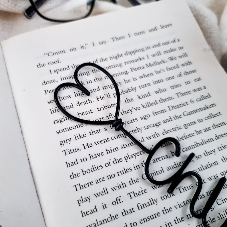 May include: A black metal heart-shaped bookmark rests on the open pages of a book. The bookmark is made of a single wire bent into a heart shape, with the word "CH" at the bottom. The book's text is visible.