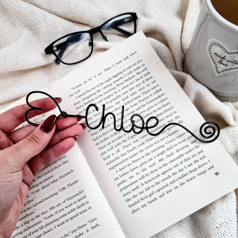 May include: A black wire bookmark in the shape of a heart and the name "Chloe" in cursive. The bookmark is resting on an open book with text. A pair of black glasses and a gray mug are in the background.